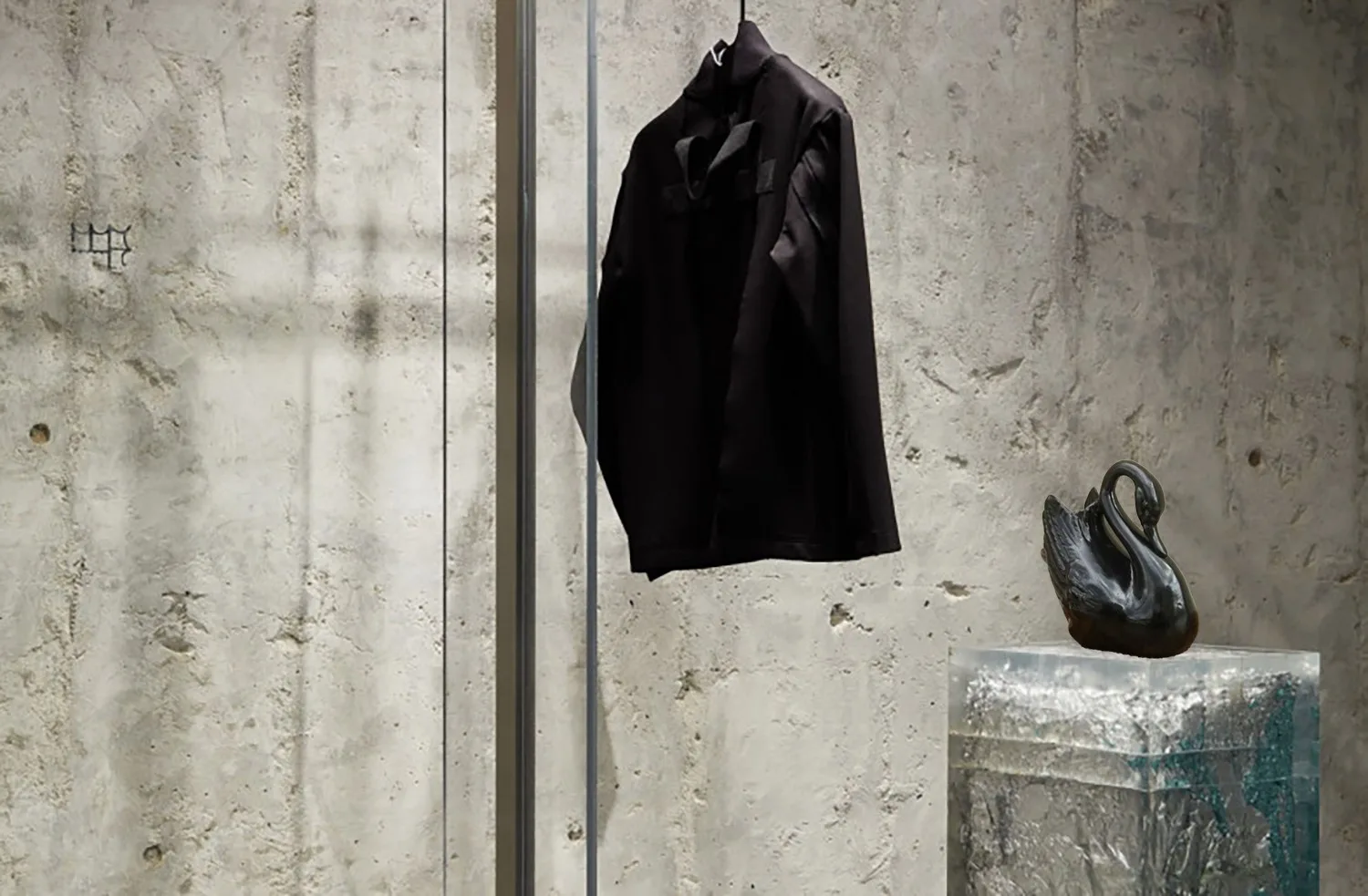 Chaqueta negra colgada y cisne negro sobre peana de resina ante una pared de hormigón, en MATERIA